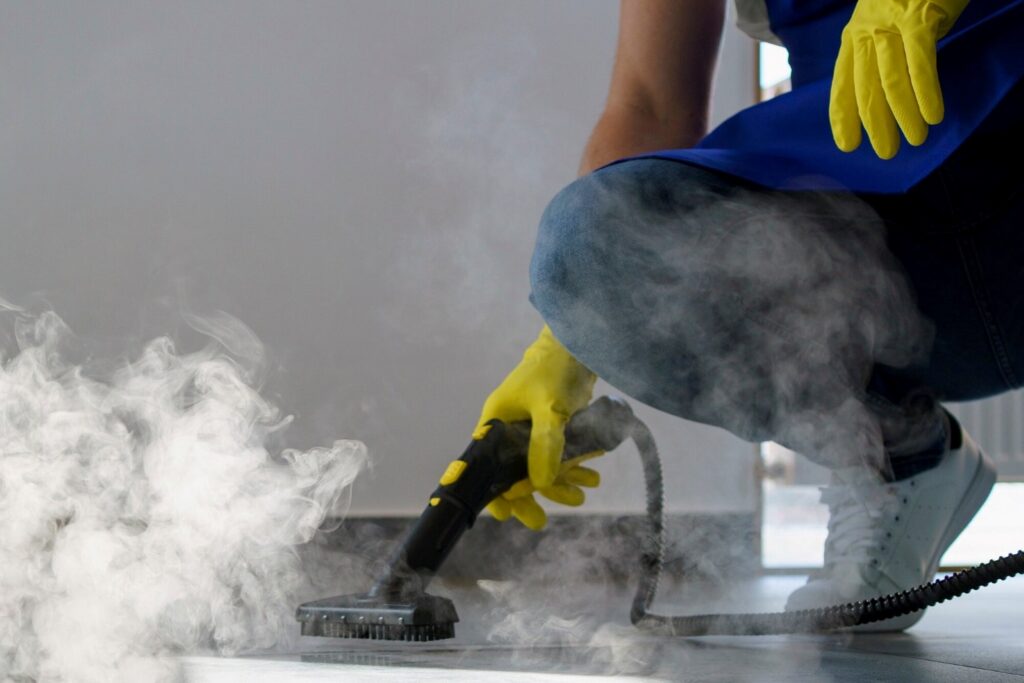 Person in yellow gloves kneels and uses a steam floor cleaner to scrub a surface, with visible steam surrounding the area.