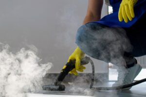 Industrial Cleaning Services 2 - CleanPro of America - CleanPro of America Person in yellow gloves kneels and uses a steam floor cleaner to scrub a surface, with visible steam surrounding the area.