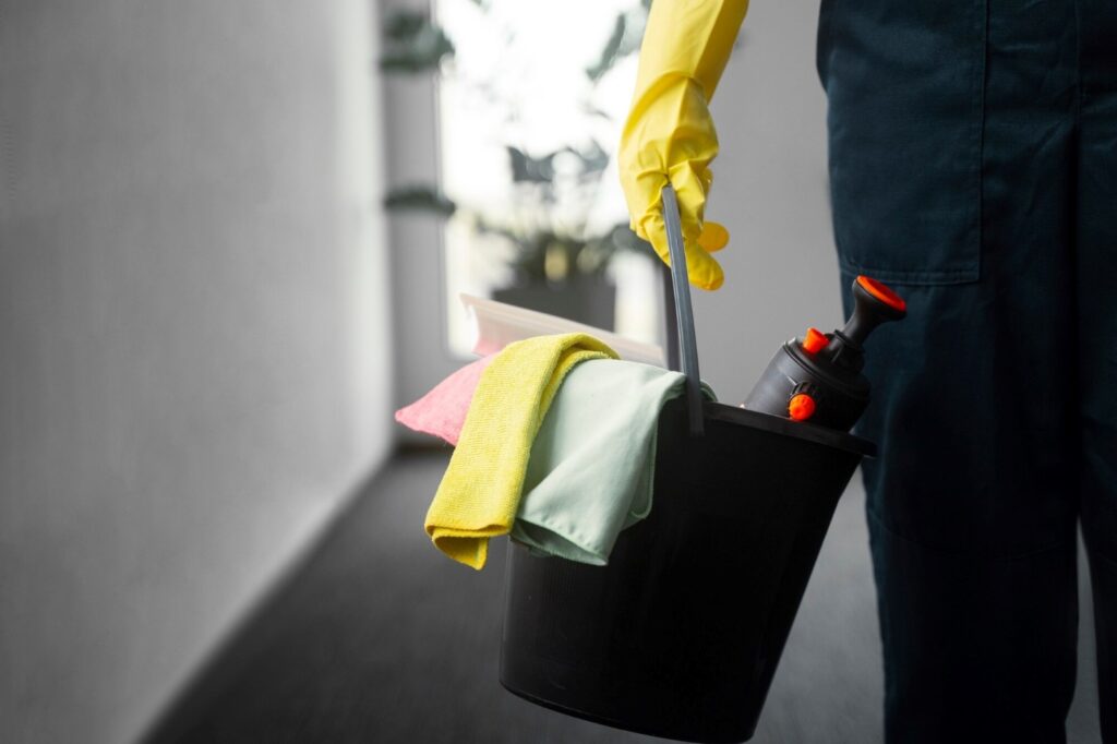 Industrial Cleaning Services 3 - CleanPro of America A cleaning worker in yellow gloves holding a black bucket with cleaning supplies (cloths, spray bottle, and a mop handle).